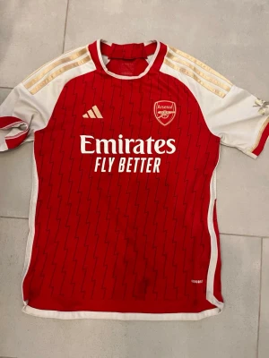 Arsenal matchtröja Adidas röd/vit - Snygg Arsenal matchtröja från Adidas i rött och vitt med gulddetaljer på axlarna. Tröjan har klubbmärke och Adidas-logga på bröstet samt 'Emirates Fly Better' tryckt framtill. Tillverkad i lätt och ventilerande material, perfekt för fotbollsträning eller match.