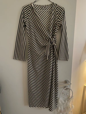 Randig omlottklänning med knyt - Snygg långärmad omlottklänning med svartvita diagonala ränder och knyt i midjan. Klänningen har v-ringning och är tillverkad i mjukt, stretchigt material som ger en bekväm passform. Perfekt för dig som gillar stilrena och trendiga plagg.