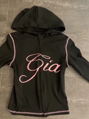 Svart loungewear-set från I.Am.Gia - Säljer ett svart loungewear-set från I.Am.Gia i Stl M med rosa sömmar och tryck. Setet består av en hoodie med dragkedja och huva samt matchande byxor med texten 'I Am Gia' i rosa på baksidan. Materialet är mjukt och stretchigt