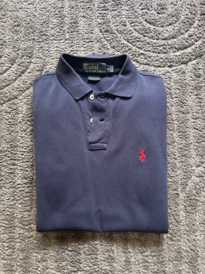 Marinblå långärmad pikétröja från Polo Ralph Lauren - Marinblå långärmad pikétröja från Polo Ralph Lauren i slim fit. Klassisk krage, två knappar och röd broderad logga på bröstet. Tillverkad i bomull för en bekväm och stilren känsla.
