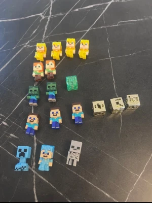 Minecraft minifigurer och block - Samling med små Minecraft-figurer i plast, inklusive karaktärer som Steve, Alex, zombies, creeper, skelett och block. Figurerna är färgglada och fyrkantiga, typiska för Minecrafts pixelstil. Perfekt för dig som gillar att samla eller leka med Minecraft-prylar.