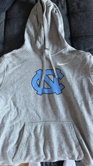 Grå collage Nike hoodie - Säljer en grå hoodie från Nike med stor blå logga på bröstet och vit Nike-swoosh på sidan. Tröjan har huva med dragsko och en stor magficka. Tillverkad i mjukt material, perfekt för chill dagar eller när du vill ha en sportig look.