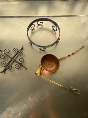 Sats vintage köksredskap i metall - Säljer ett set med tre vintage köksredskap i metall: en svart grytunderlägg med snirkligt mönster, en rund ställning i svart metall samt en kopparfärgad mässings skopa med tvinnat handtag. Perfekt för dig som gillar retrostil och unika detaljer i köket.