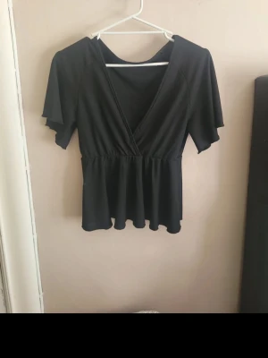 Svart v-ringad blus med volang - Svart blus med v-ringning och korta ärmar. Toppen har en omlott-look framtill och volang nertill som ger en snygg siluett. Perfekt att styla med jeans eller kjol för en trendig look.