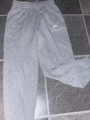 Gråa mjukisbyxor från Nike - Säljer ett par klassiska gråa mjukisbyxor från Nike med resår i midjan och muddar vid bensluten. Byxorna har en enkel och clean design med Nike-logga broderad på vänster ben. Perfekta för chill dagar eller träning.
