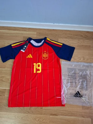 Spanien fotbollströja Adidas Lamine yamal #19 VM 26/27 - Röd och blå Spanien fotbollströja från Adidas med gula detaljer, nummer 19 och Lamine Yamal på ryggen. Tröjan har korta ärmar, tunnt funktionsmaterial och klassiska ränder. Perfekt för dig som älskar fotboll och vill stötta landslaget.