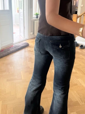 Mörkblå bootcut jeans med fickdetalj - Snygga mörkblå bootcut jeans med lätt slitning och dekorativ knapp på bakfickan. Jeansen har klassisk femficksdesign och är tillverkade i denim med stretch för skön passform. Perfekta för en trendig och avslappnad look. Inerbenslängd = 82 cm. Midja = 41 cm rakt över