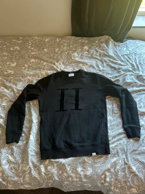Svart sweatshirt från Les Deux - Stilren svart sweatshirt från Les Deux med ett stort, svart sammetsliknande 'II'-tryck på bröstet. Tröjan har rund halsringning, ribbade muddar vid ärmslut och nederkant samt en mjuk insida. Perfekt för en clean och modern look. Lite slitningar där fram.