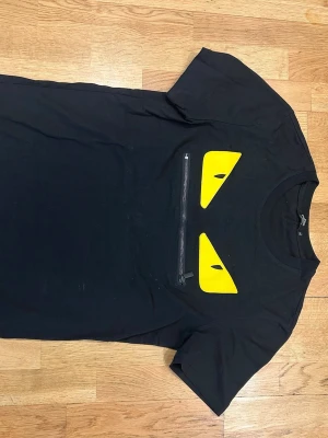 Svart Fendi t-shirt  - Cool svart Fendi t-shirt med två gula ögonliknande detaljer och en dekorativ dragkedja på bröstet. T-shirten har rund hals och korta ärmar. Perfekt för dig som vill sticka ut med en unik och edgy stil.