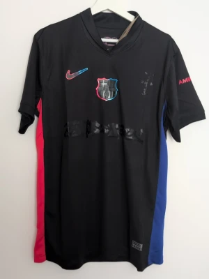 Svart FC Barcelona X Travis Scott  - Säljer en svart FC Barcelona matchtröja från Nike med klubbmärke och Nike-logga i neonfärger på bröstet. Tröjan har röda och blå sidopaneler, korta ärmar och UNHCR-tryck på ryggen. Perfekt för fotbollsträning eller att bära som supporter.