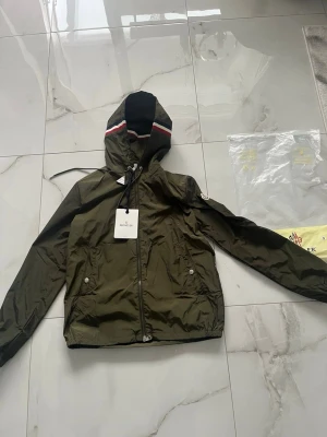 Olivgrön vindjacka från Moncler - Snygg olivgrön vindjacka från Moncler med huva och dragkedja framtill. Jackan har två fickor med tryckknappar och en diskret logga på ärmen. Detaljer med röd och vit rand på huvan ger en sportig touch. Tillverkad i lätt, glansigt material.