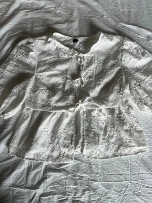 Vit blus med broderade blommor - Söt vit blus med broderade blommor och knytning framtill. Blusen har volang nedtill och puffiga långärmar med resår vid ärmslut. ❤️