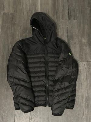 Svart pufferjacka från Hugo Boss - Pris kan diskuteras. Snygg svart pufferjacka från Hugo Boss med huva och dragkedja framtill. Jackan har quiltad design, diskret logga på ärmen och bak på ryggen samt elastiska muddar. Perfekt för kalla dagar och riktigt trendig look.