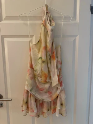 Blommig halterneck kortklänning - Säljer en söt kortklänning med halterneck och volangkant. Klänningen har ett blommigt mönster i ljusa toner av rosa, grön, gul och orange. Materialet känns lätt och luftigt, perfekt för varma dagar. Smock i ryggen för skön passform och omlottliknande fall framtill.