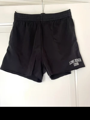 Mörkgråa shorts  - Mörkgråa shorts från H&M Divided med trycket 'LONG BEACH 1998' i vitt på ena benet. De har elastisk midja och är tillverkade i en mjuk bomullsblandning, perfekta för avslappnade dagar.
