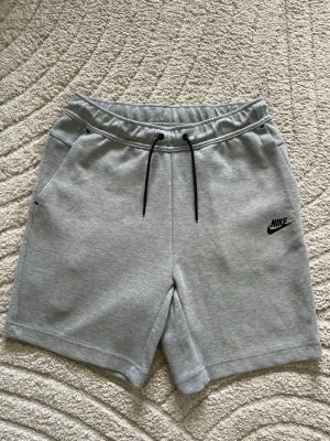Nike tech shorts grå - Snygga Grå sweatshorts från Nike med svart logga på vänster ben. Shortsen är endast använda 2 gånger i nyskick , beställningsbekräftelse finns om så önskas efter köp!