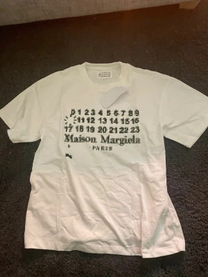 Vit Maison Margiela t-shirt - Vit t-shirt från Maison Margiela med svart tryck framtill som visar siffror och logotypen. Klassisk passform med rund hals och korta ärmar. Perfekt för dig som gillar stilrena och trendiga plagg med en twist.