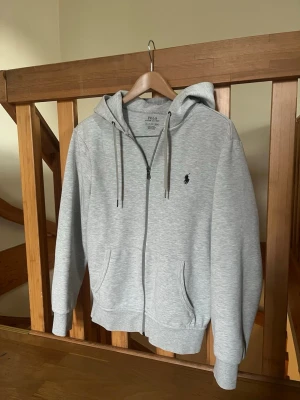 Ljusgrå hoodie från Polo Ralph Lauren - Snygg ljusgrå hoodie med dragkedja från Polo Ralph Lauren. Klassisk design med huva och dragsko, två fickor framtill och broderad logga på bröstet. Perfekt för en avslappnad och stilren look. 
