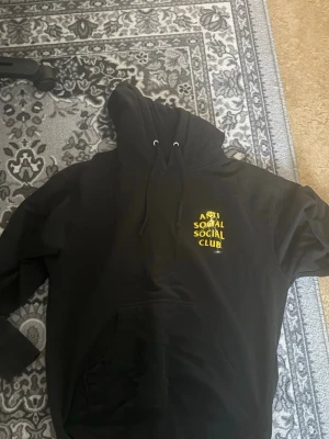 Svart hoodie Anti Social Social Club - Svart hoodie med stor gul Anti Social Social Club-logga på ryggen och mindre logga på bröstet. Klassisk känguruficka framtill och justerbar huva med snören. Tillverkad i mjukt material, perfekt för en avslappnad streetwear-look.