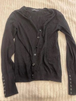 Svart kofta från Gina Tricot - Svart tunn kofta från Gina Tricot med knappar framtill och dekorativa knappar vid ärmsluten. Långärmad och i mjukt, rätt så stretchigt material. 