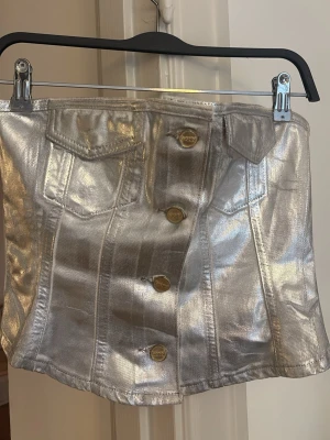 Kort silver topp med knappar - Snygg kort kjol i metallic-silver med markerade sömmar och dekorativa fickor framtill. Kjolen har en rak passform och stängs med stora knappar framtill. Perfekt för dig som vill sticka ut med en trendig look.