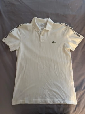 Vit pikétröja från Lacoste - Klassisk vit pikétröja från Lacoste med krage och knappar framtill. Tröjan har korta ärmar med band längs axlarna där det står 'Lacoste' och den ikoniska gröna krokodilloggan på bröstet. Regular fit och tillverkad i bomull.