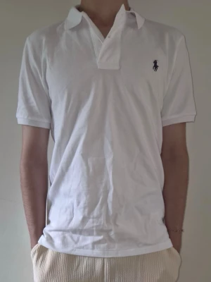 Vit pikétröja med Ralph lauren logga - Klassisk vit pikétröja med Polo Ralph Lauren. Korta ärmar och den ikoniska loggan broderad på bröstet. Tröjan har krage och knappar vid halsen. Perfekt till jeans eller shorts för en clean och stilren look.