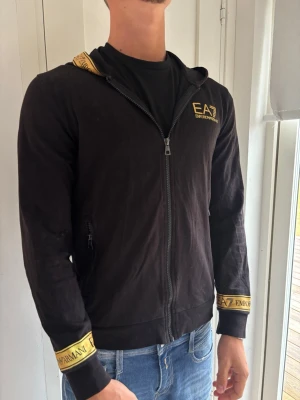 Svart EA7 hoodie med dragkedja - Snygg svart hoodie från EA7 Emporio Armani med gul logga på bröstet och detaljer på huva och ärmslut. Dragkedja framtill och två fickor med dragkedja. Perfekt för dig som vill ha en stilren och sportig look.