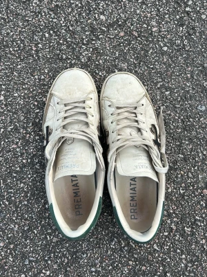 Vita Premiata sneakers med grön detalj - Säljer ett par vita sneakers från Premiata med svart logga på sidan och grön detalj på hälen. Skorna har klassisk låg profil, vita snören och ovandel i läder. Perfekta för dig som gillar stilrena och tidlösa sneakers.