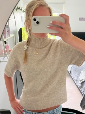Beige stickad t-shirt med polokrage - Kortärmad stickad t-shirt i mjuk beige färg med hög polokrage. Tröjan har en rak passform och är tillverkad i ett tunt, mysigt material som passar perfekt till jeans eller kjol. Enkel och stilren design utan synligt märke.