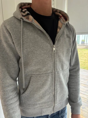 Burberry Grå zip hoodie med mönstrad huva - Snygg grå hoodie med dragkedja och fickor framtill. Huvan har ett klassiskt rutigt mönster på insidan och justerbara snören. Tillverkad i mjukt sweatshirtmaterial, perfekt för en avslappnad stil.