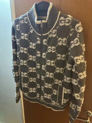 Grå stickad ziptröja från Gucci - Snygg grå stickad ziptröja från Gucci med vit GG-logga i mönstret. Tröjan har hög ribbad krage, dragkedja framtill och fickor på sidorna. Detaljer i vitt vid krage och muddar. Perfekt för dig som gillar exklusiva streetwear-vibbar.