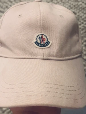 Ljusrosa keps från Moncler - Säljer en stilren ljusrosa keps från Moncler med klassisk böjd skärm och broderad Moncler-logga framtill. Kepsen är tillverkad i bomullstyg och har justerbar passform baktill. Perfekt accessoar för en trendig och avslappnad look.