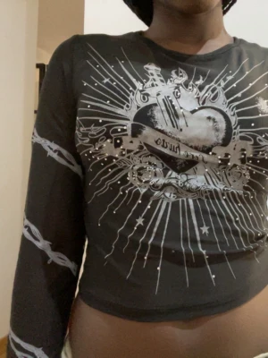 Emo topp - Snygg svart croppad topp med lång ärm och silverfärgat tryck med hjärta och detaljer framtill. Toppen har coola grafiska mönster på ärmarna och små glittriga detaljer som ger extra edge. Perfekt för dig som gillar statement-plagg. DEN ÄR LITE GENOMSKINLIG 