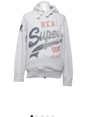 Ljusgrå hoodie från Superdry - Ljusgrå hoodie från Superdry med stor tryckt logga i marinblått och rött framtill. Huva med dragsko och ribbade muddar vid ärmar och nederkant. Tryck även på ärmen och ryggen. Perfekt för en avslappnad streetwear-look.