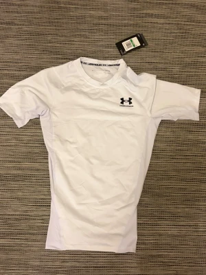 Vit kompressionströja Under Armour - Säljer en vit kompressionströja från Under Armour med korta ärmar och rund hals. Tröjan har en tight passform och är tillverkad i ett stretchigt funktionsmaterial som andas, perfekt för träning och sportaktiviteter. Diskret logga på bröstet.