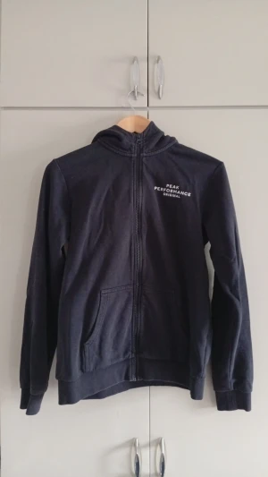 Kofta Peak Performance - Snygg mörkblå hoodie från Peak Performance med dragkedja framtill och två fickor. Klassisk logga på bröstet och skön huva. Tillverkad i mjukt bomullsmaterial som känns bekvämt hela dagen. Perfekt för en avslappnad och sportig stil.