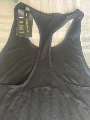 Svart Nike Dri-FIT träningstopp - Snygg svart träningstopp från Nike med Dri-FIT-teknologi som håller dig torr under löpning eller gympass. Modellen har brottarrygg och reflekterande Nike-logga framtill. Tillverkad i ett stretchigt och ventilerande material för maximal komfort.