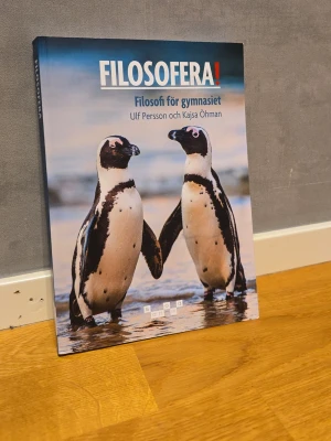 Filosofibok för gymnasiet - Filosofiboken 'Filosofiera! Filosofi för gymnasiet' av Ulf Persson och Kajsa Öhman. Omslaget har två pingviner mot en strandbakgrund. Boken är i mjukpärm och har blå och vita färgtoner.