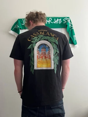 Svart Avenida Casablanca t-shirt - Svart t-shirt med färgglad tryckt grafik på både bröstet och ryggen, där det står 'Avenida Casablanca' och ett fönster med gröna blad och byggnadsmotiv. Klassisk passform och korta ärmar. Perfekt för dig som gillar unika prints och streetwear vibes. Pris går även att diskutera!
