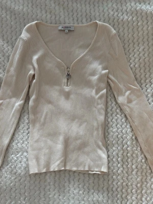 Beige ribbad topp med dragkedja - Snygg beige ribbad topp från Norgani med v-ringning och dekorativ dragkedja framtill. Långärmad modell i mjukt material som sitter figurnära. Perfekt att styla med jeans eller kjol för en trendig look.
