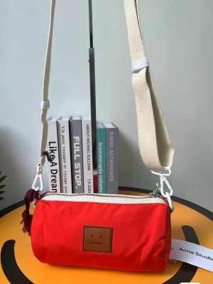 Röd axelväska Acne Studios - Cool röd axelväska från Acne Studios med beige justerbar axelrem och vit dragkedja. Väskan har en cylinderformad design, dekorativ tofs och Acne Studios klassiska ansiktslogga framtill. Perfekt för att bära det viktigaste med stil.