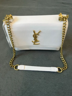 Vit YSL handväska med guldkedja - Snygg vit handväska från YSL med guldfärgad kedjerem och ikonisk YSL-logga framtill. Väskan är i slätt läder och har en stilren, rektangulär form. Perfekt accessoar för att lyfta vilken outfit som helst.