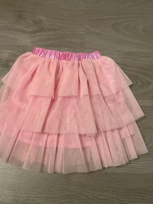Rosa volangkjol med tyll - Supersöt rosa kjol med flera lager tyll och volanger. Kjolen har ett glansigt resårband i midjan som ger en extra fin detalj. Perfekt för dig som gillar en lekfull och fluffig stil.