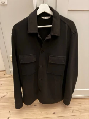 Svart overshirt från Zara - Stilren svart overshirt från Zara med klassisk krage och två stora bröstfickor med lock. Skjortjackan har raka ärmar och knäppning framtill. Perfekt lager-på-lager-plagg för en trendig look.