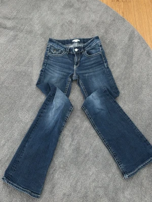 Mörkblå bootcut jeans med slitningar - Säljer ett par mörkblå bootcut jeans med snygga slitningar och klassisk femficksmodell. Jeansen har låg midja och utsvängda ben, vilket ger en retro och trendig look. Perfekta för dig som gillar en avslappnad men ändå stilsäker stil.