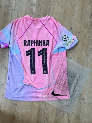Barça matchtröja Raphinha rosa/lila - Säljer en FC Barcelona matchtröja från Nike i rosa och lila toner med blå detaljer. Tröjan har Raphinha och nummer 11 på ryggen, Champions League-märke på ärmen och Spotify-logga på bröstet. Perfekt för dig som älskar fotboll och vill sticka ut på planen.
