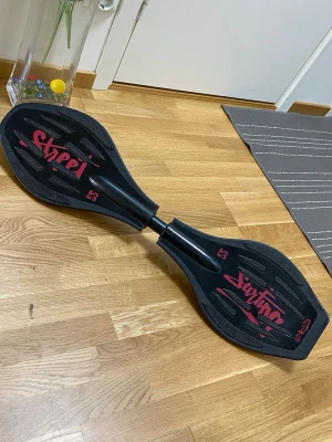 Street Surfing Waveboard   - Cool svart waveboard med röd text och detaljer. Brädan har två plattor som sitter ihop med en metallstav i mitten och är designad för att svänga smidigt på asfalt. Perfekt för dig som gillar att cruisa runt och testa tricks. Längden är ca 85cm 