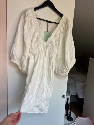 Vit kort omlottklänning med puffärm - Supersöt vit kort klänning med omlottdesign och puffiga trekvartsärmar. Klänningen har djup v-ringning och fina volangdetaljer längs ringningen. Perfekt för sommarens alla tillfällen.