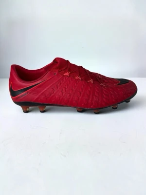 Röda Nike Hypervenom fotbollsskor - Säljer ett par röda Nike Hypervenom fotbollsskor med svart sula och svarta detaljer. Skorna har en modern, låg profil och är tillverkade i syntetmaterial med struktur för bättre bollkontroll. Perfekta för spel på gräsplaner och har klassisk snörning. Pris kan diskuteras